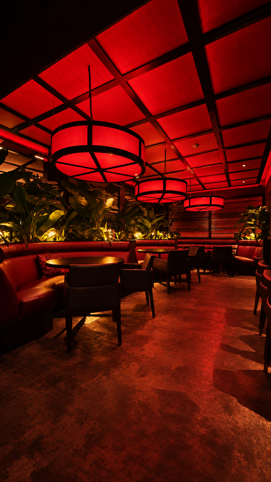 Dining room after dark, large glowing lanterns hanging beneath a red-lit gridded ceiling above banquettes and tropical planting.