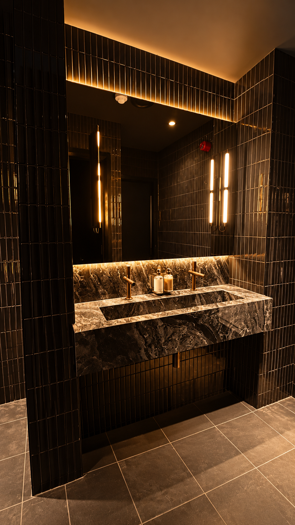 Black and gold marble basin with bronze taps beneath tube pendants and an amber cove glow.
